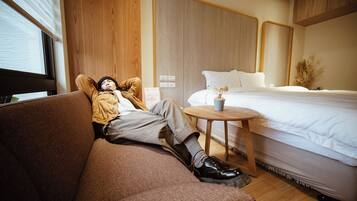 Habitación de hotel con pared de paneles de madera, cama con ropa blanca, mesita de madera redonda con un florero, y una persona acostada en un sofá marrón.