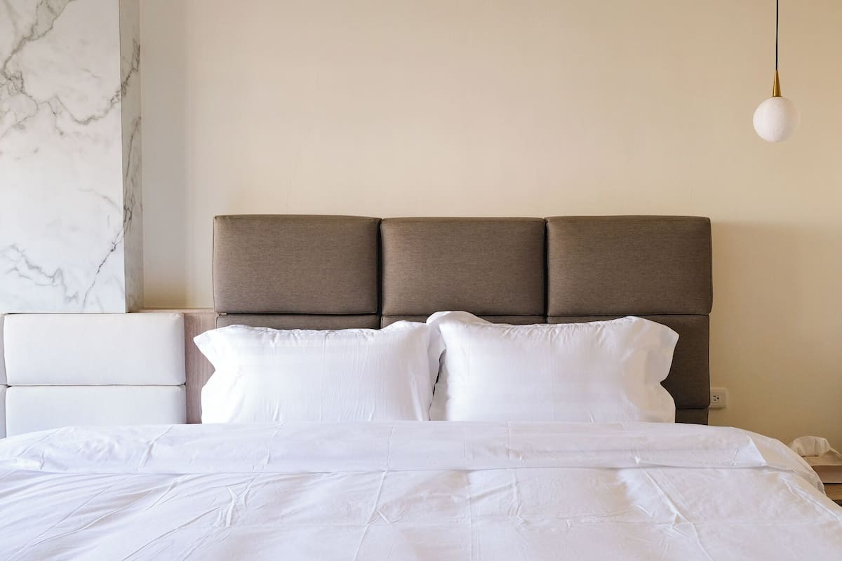 A neatly made bed with a headboard, a white nightstand, and a marble wall.