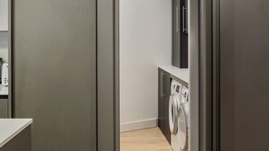 Een moderne wasserij met een wasmachine en droger, houten vloeren en een keuken die zichtbaar is door een doorgang.