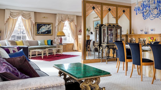 Ein geräumiges Wohnzimmer mit einem grünen Couchtisch, einer Couch mit bunten Kissen, einem Essbereich mit blauen Stühlen und einem großen Spiegel, der den Raum reflektiert.