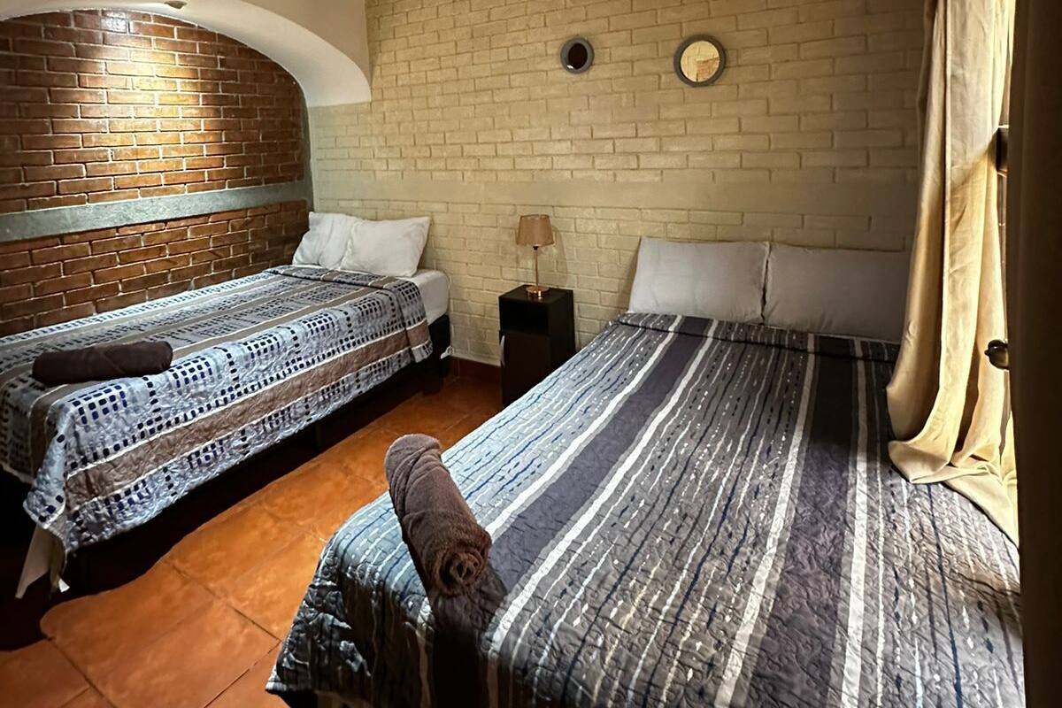 Una habitación con dos camas, un muro de ladrillo, techo de madera y suelo de baldosas.