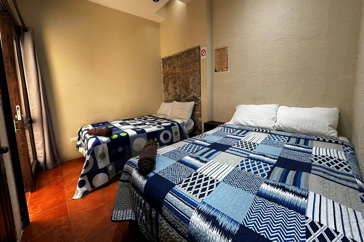 Habitación con dos camas, cada una con ropa de cama estampada. Se ve una puerta a la izquierda y una pared con textura y una pequeña señal.
