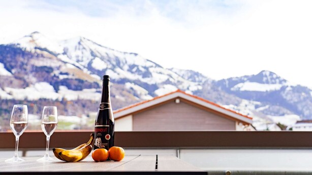 A bottle of Heidenblut wine, two glasses, a banana, and two oranges on a balcony with a mountain view.