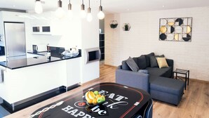 A modern kitchen with a black countertop, a dining table with a fruit bowl, and a grey sofa in a living area.