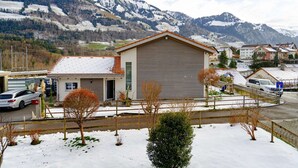 A single-story building with a garage, surrounded by snow-covered ground and trees, with mountains in the background.