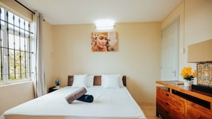 A neatly made bed with white linens and a brown towel, a wooden dresser with a mirror, and a painting on the wall.