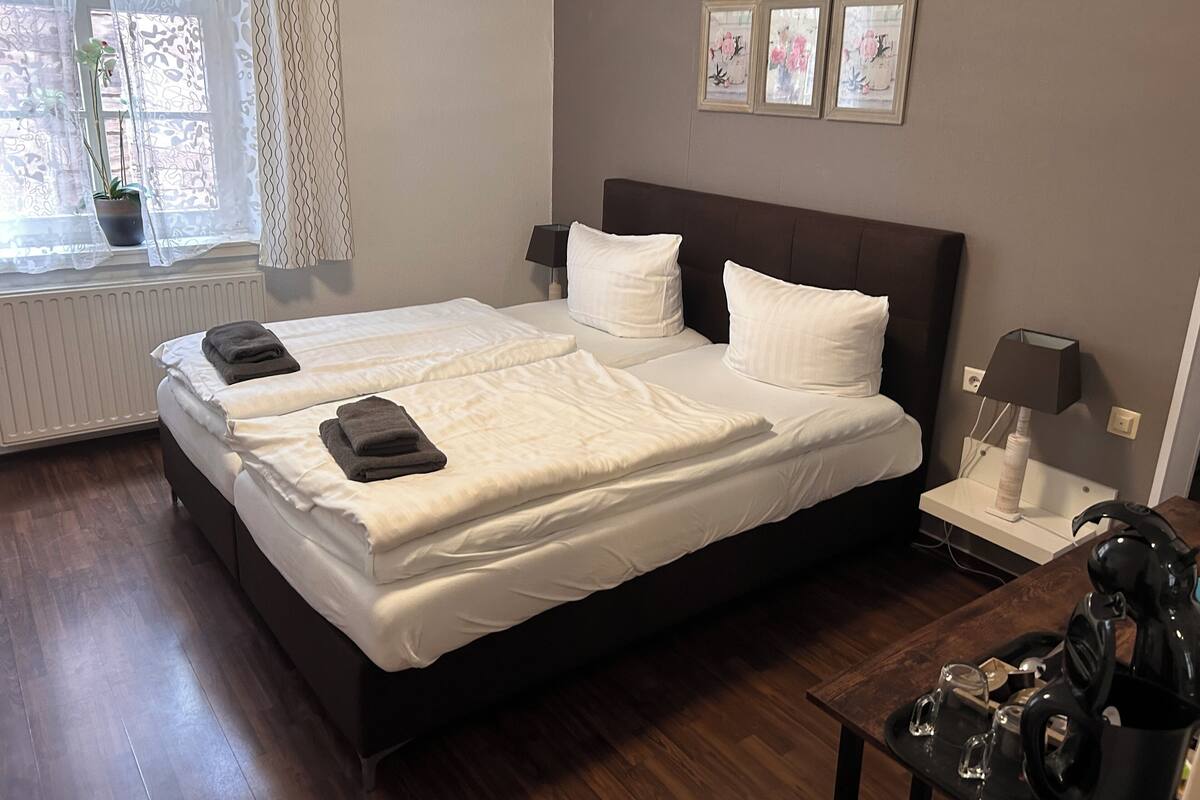 A double bed with white linens, two bedside tables with lamps, a potted plant, and a window with sheer curtains.