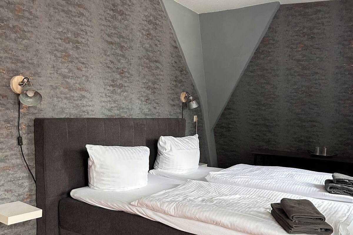 A neatly made bed with white linens and a dark headboard in a room with wooden flooring and textured gray walls.