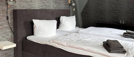 Una cama bien hecha con sábanas blancas y un cabecero oscuro en una habitación con suelo de madera y paredes grises con textura.