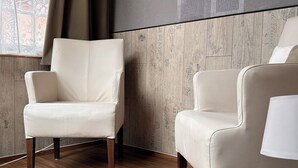 A room with two beige armchairs, wooden flooring, a large window with curtains, and a wall with a textured pattern.