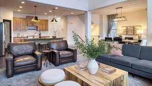 Een ruime woonkamer met lederen fauteuils, een houten salontafel en een grijze bank. Op de achtergrond is een eethoek te zien, met een keuken en een trap naar een hoger gelegen verdieping.