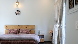 A bedroom with a wooden bed, a nightstand, a bedside table, a floor lamp, a window with curtains, and a ceiling fan. - DeerWood Cottages (Tehri)