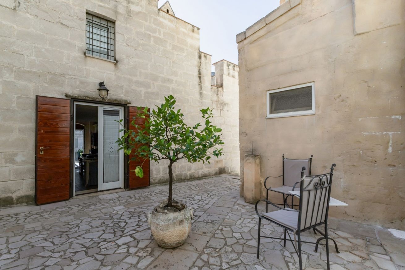 Une cour avec un sol en pierre, un arbre en pot, une chaise en métal et un bâtiment doté d’une porte et d’une fenêtre en bois.
