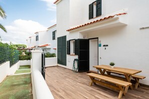Un patio con una mesa y bancos de madera, un edificio blanco con contraventanas negras y un área de jardín verde.