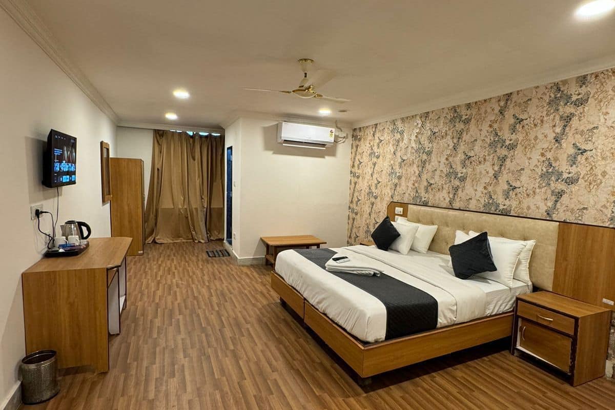 Quarto com piso de madeira, cama com roupa de cama branca e preta, mesa de cabeceira, televisão na parede e aparelho de ar condicionado.