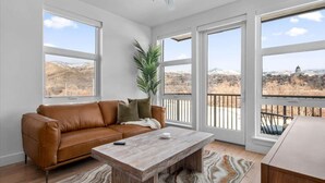 Un salón espacioso con un sofá de cuero marrón, una mesa de centro de madera y ventanas amplias que ofrecen vistas a un paisaje nevado y un edificio.