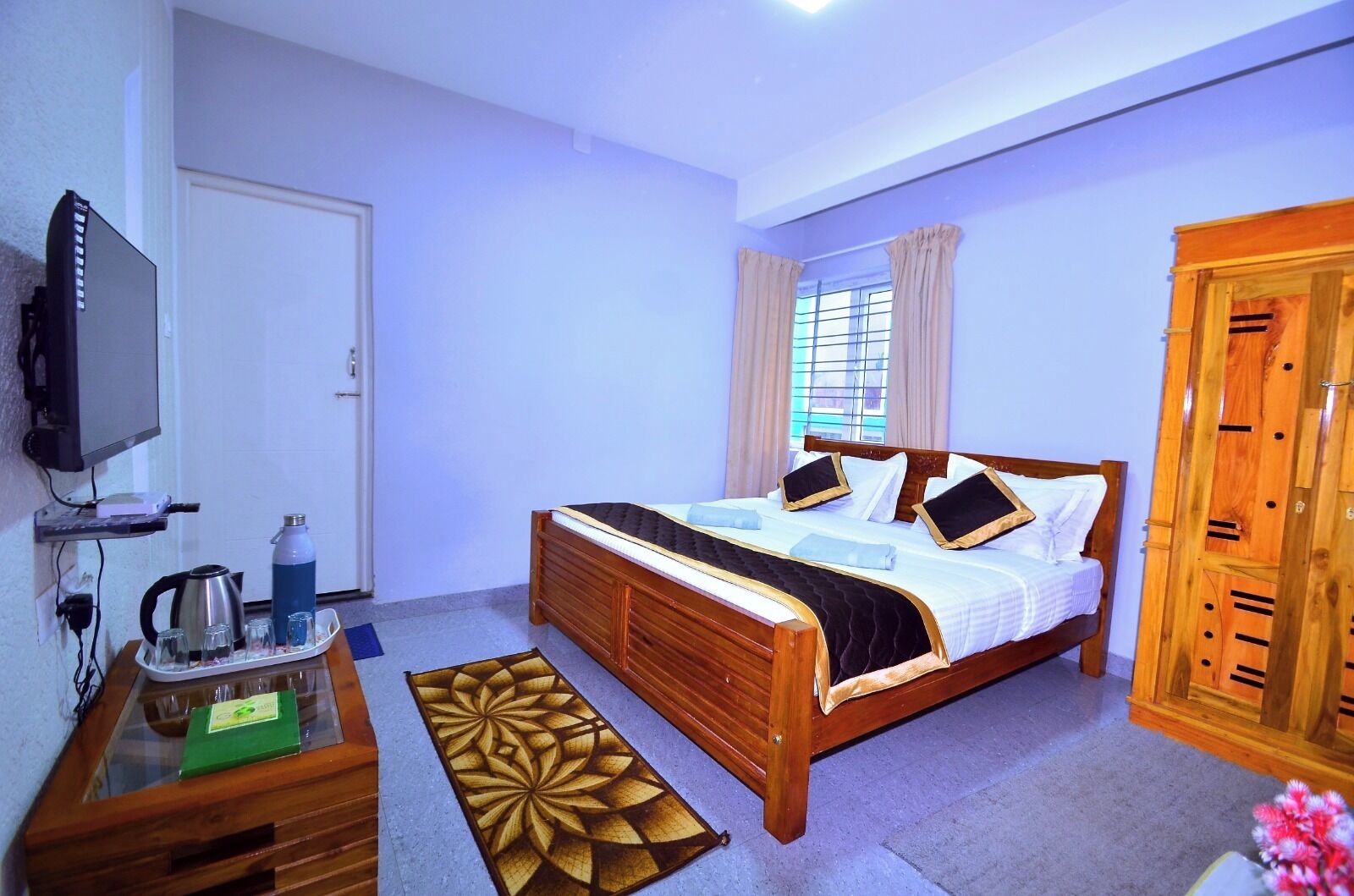 A bedroom with a wooden bed, a TV on a stand, a desk with a kettle, and a window with curtains.
