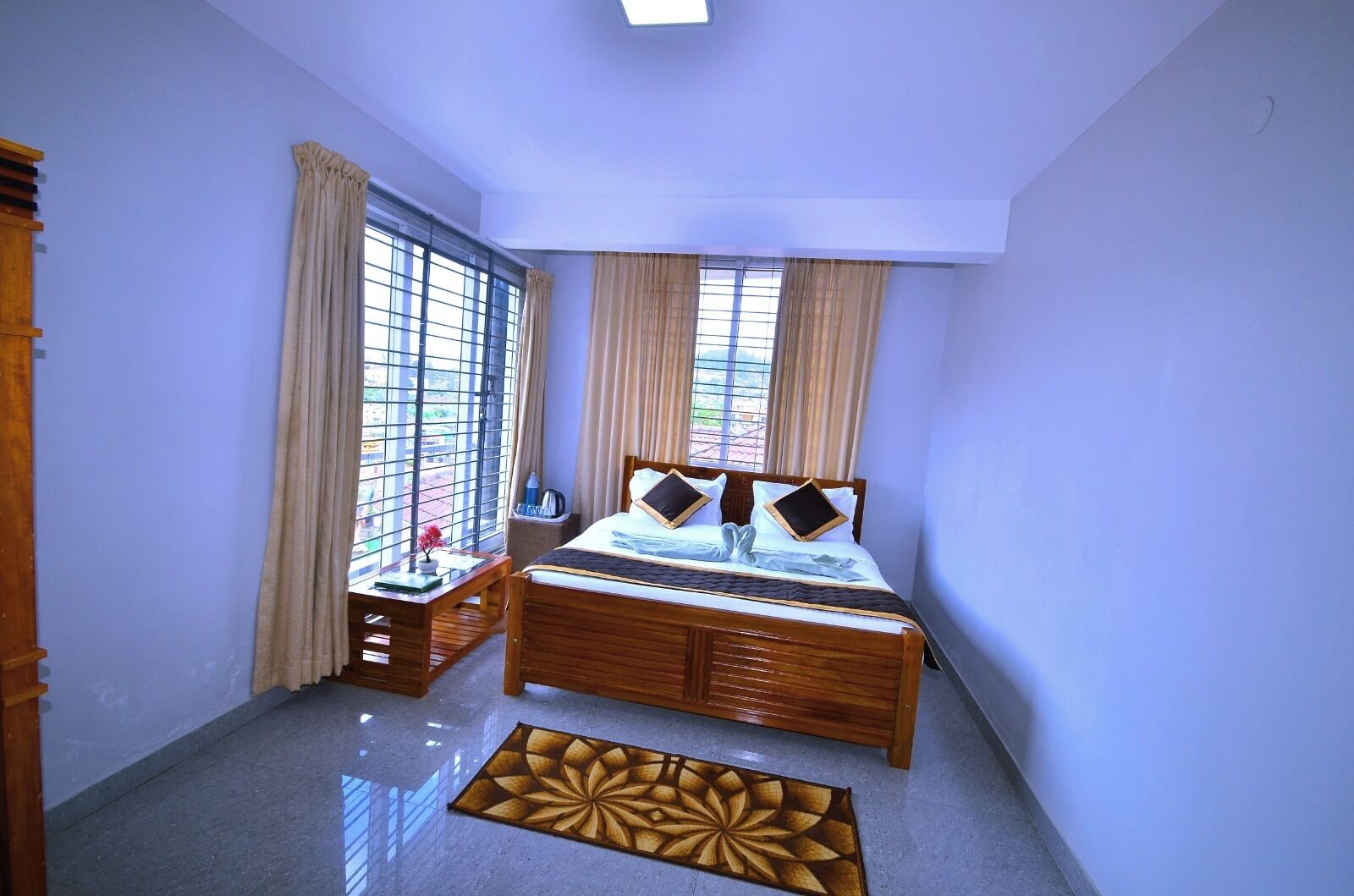 A bedroom with a wooden bed, two windows with curtains, a bedside table with a water bottle, and a decorative rug.