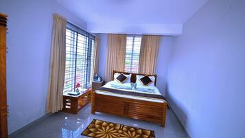 A bedroom with a wooden bed, two windows with curtains, a bedside table with a water bottle, and a decorative rug.