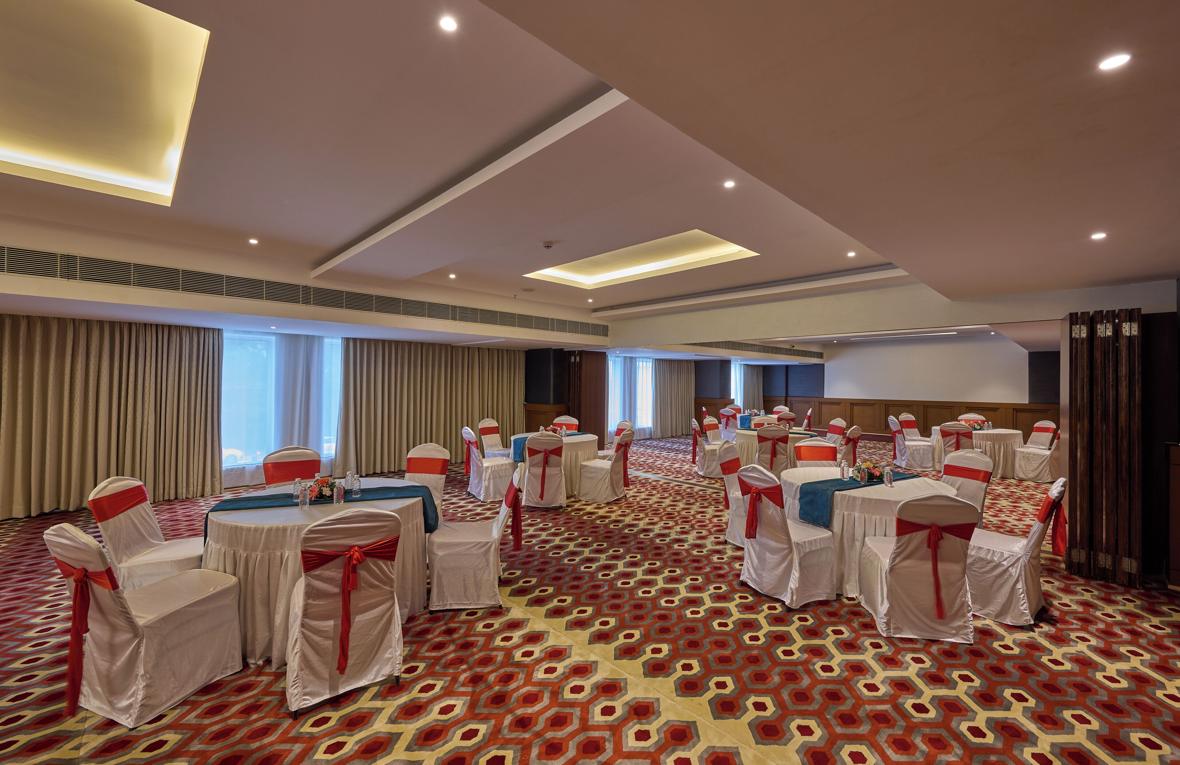 Une salle de conférence avec des tables rondes, des chaises blanches ornées de rubans rouges et un tapis à motifs.