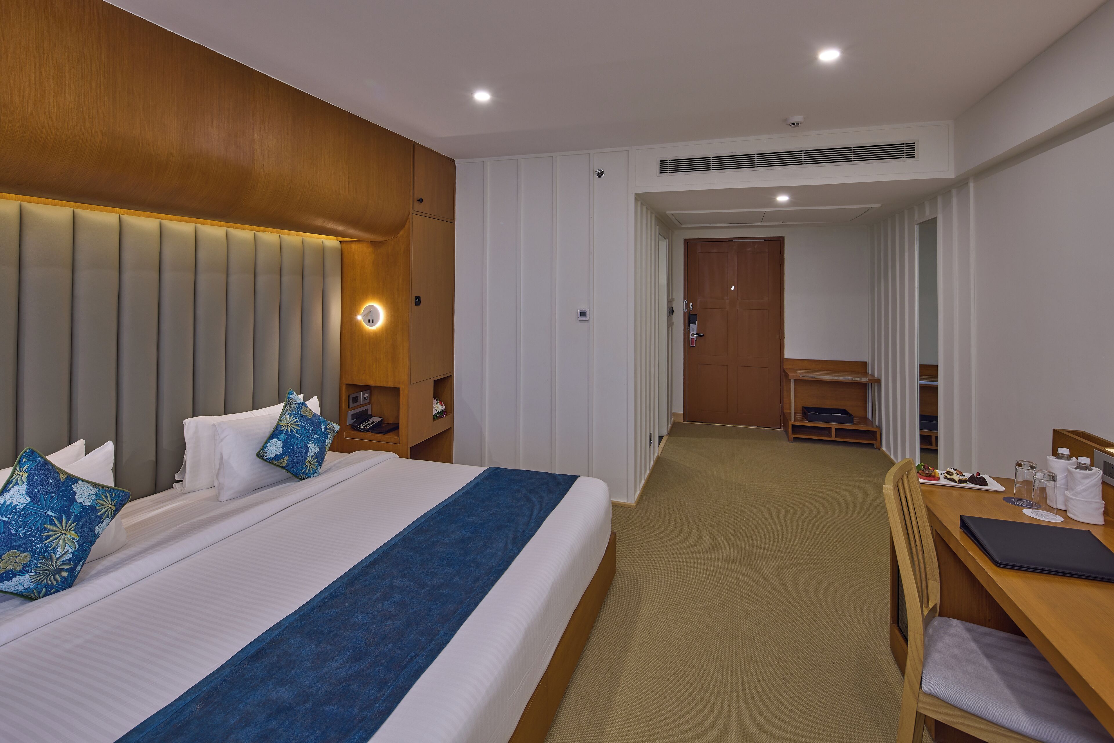 A hotel room with a large bed, a desk, and a chair. There is a wooden headboard, a bedside table, and a small shelf.