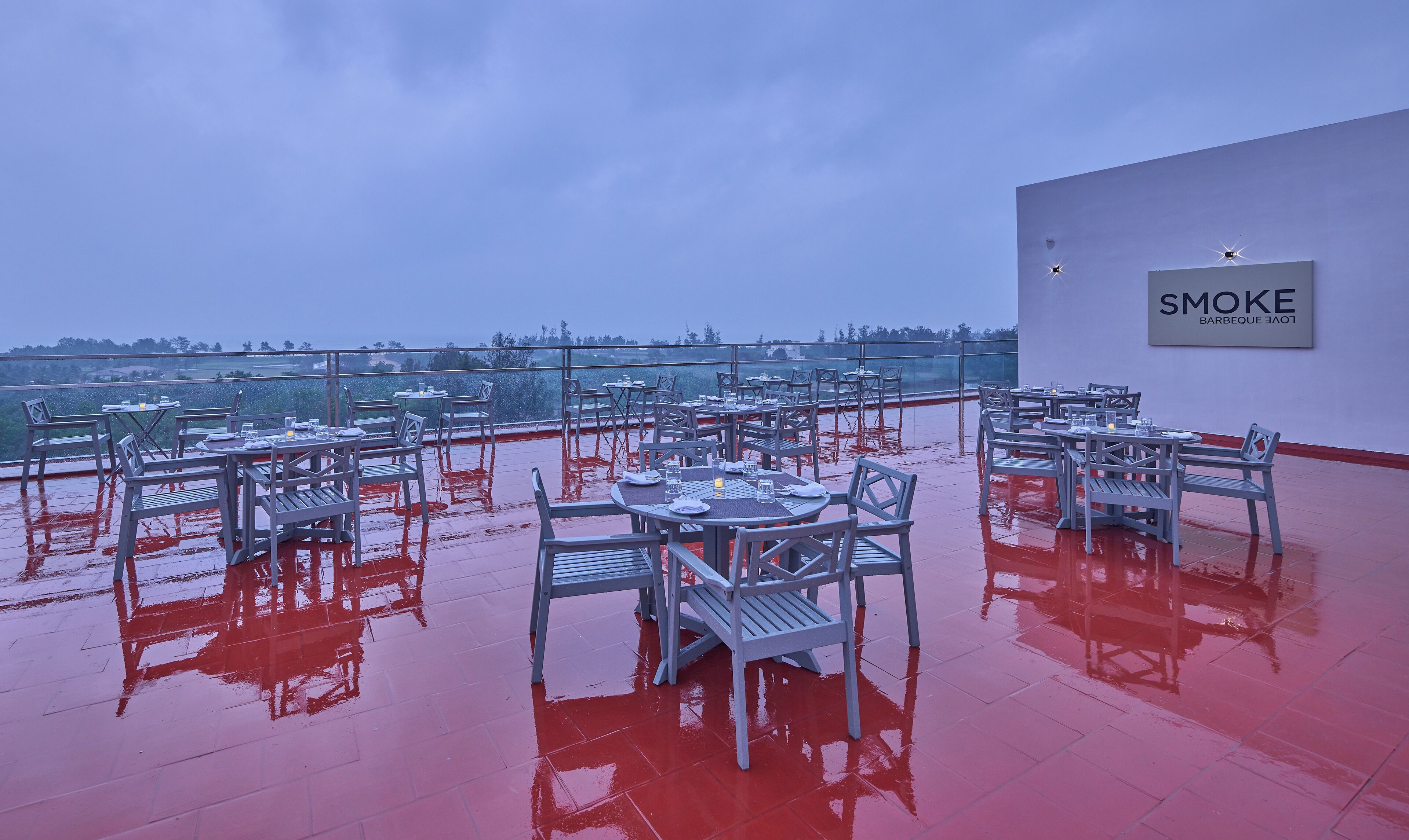 A rooftop bar with tables and chairs, a glass railing, and a sign reading "SMOKE Barbeque Lounge".