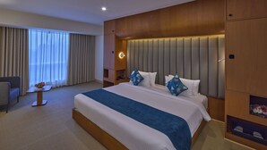 Habitación de hotel con una cama grande, una mesita de noche con un frutero, una silla y vistas al exterior a través de cortinas translúcidas.
