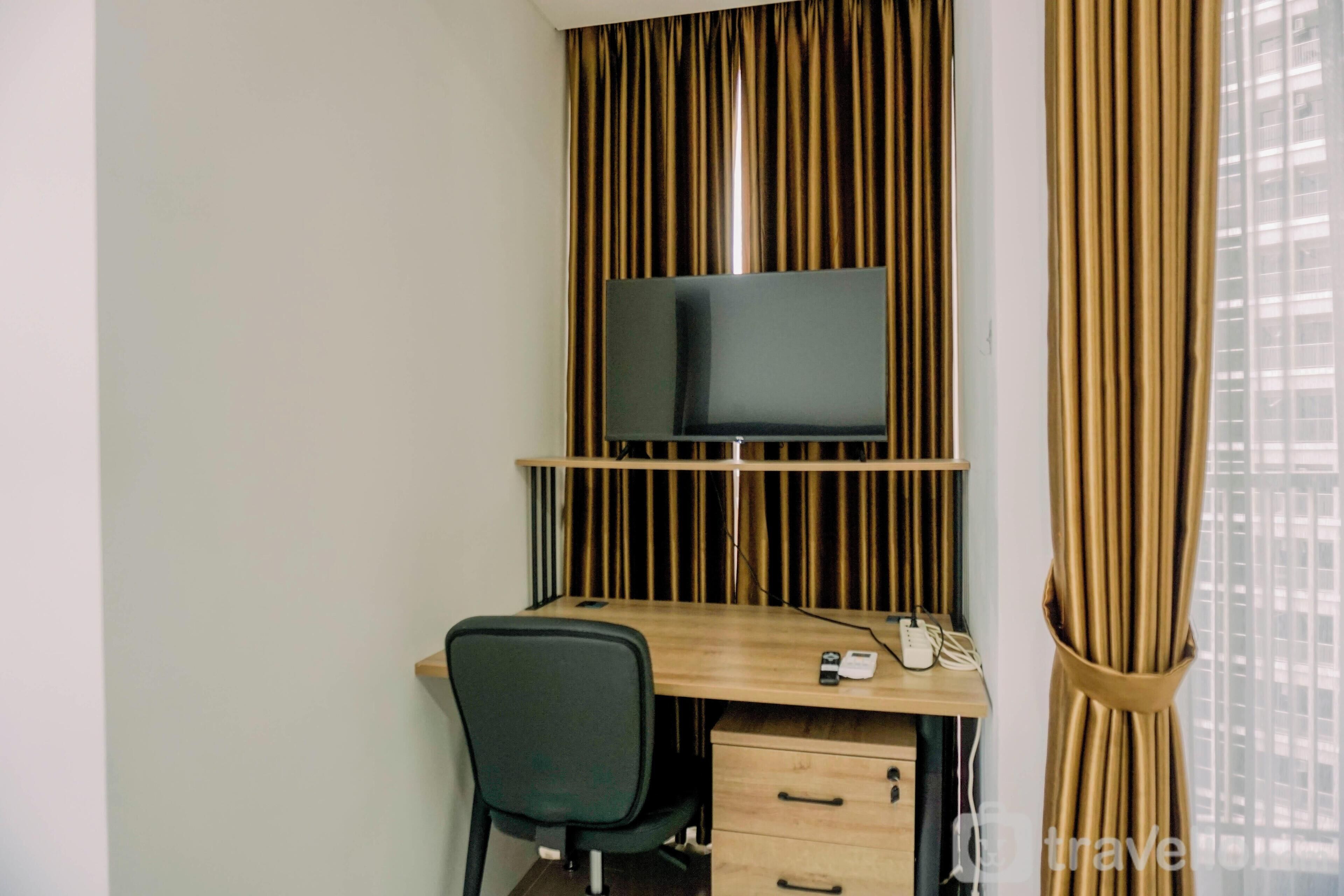A hotel room with a wooden desk, a black chair, a flat-screen TV mounted on a wooden panel, and a telephone.