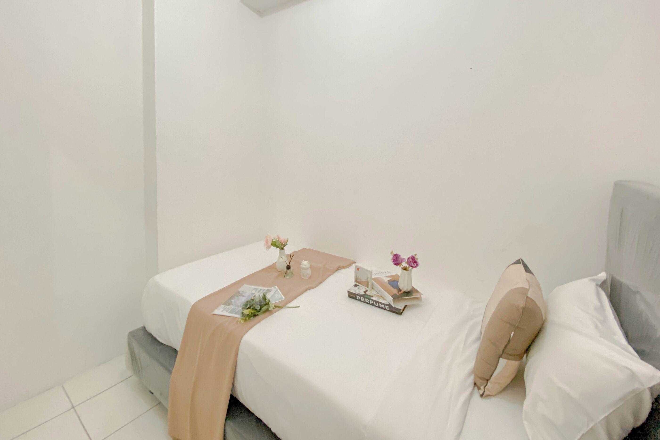 A neatly made bed with a white and beige bedspread, a small table with a magazine, a perfume box, and a vase with flowers.