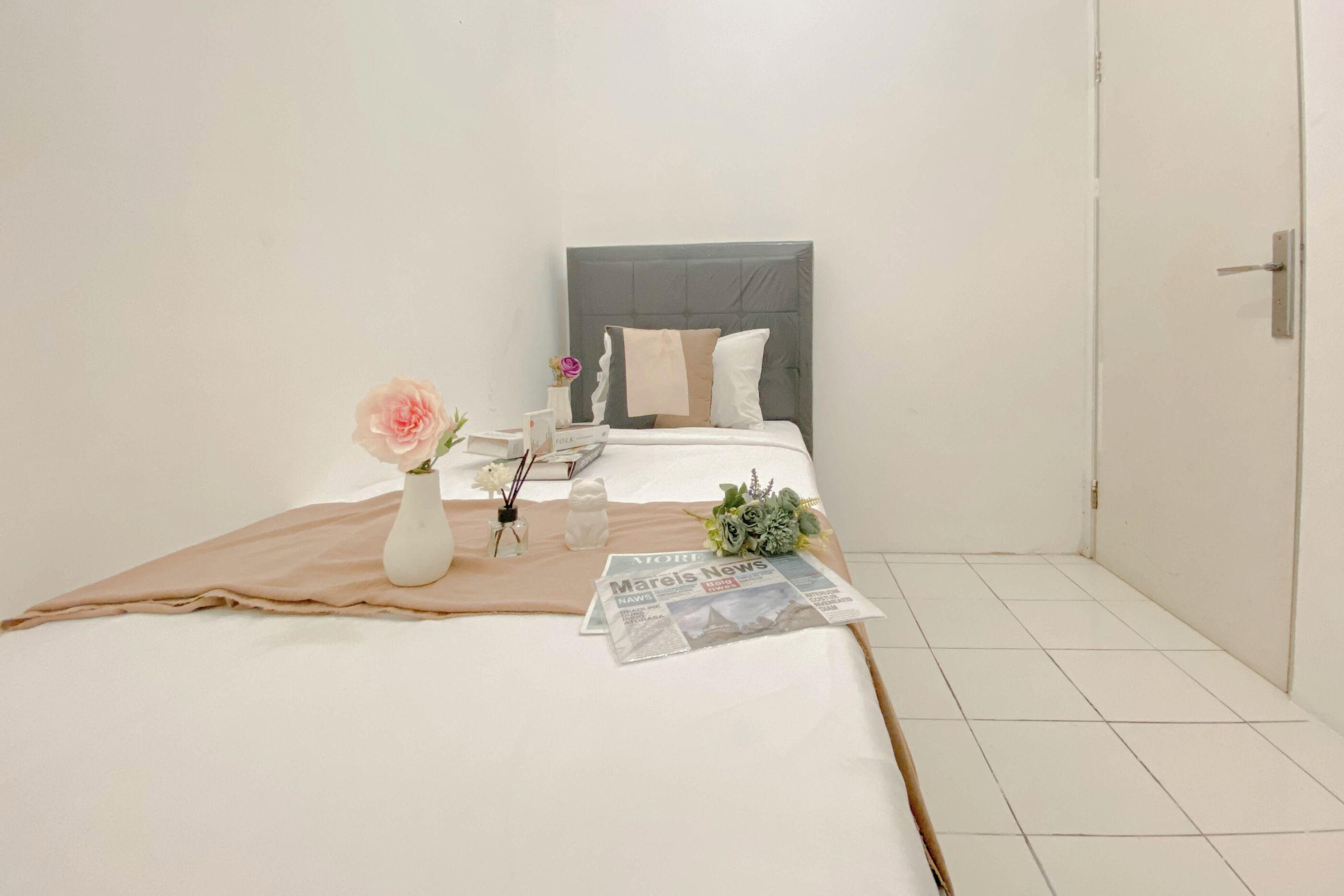 A neatly made bed with a white comforter, a beige blanket, and a bouquet of flowers. There is a small table with a vase, a bottle, and a newspaper. The room has white walls and a tiled floor.