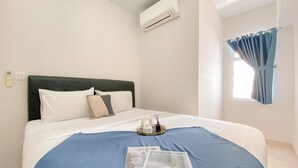 Une chambre d’hôtel avec un lit, une fenêtre avec des rideaux, un climatiseur et un plateau avec une bougie et un magazine.