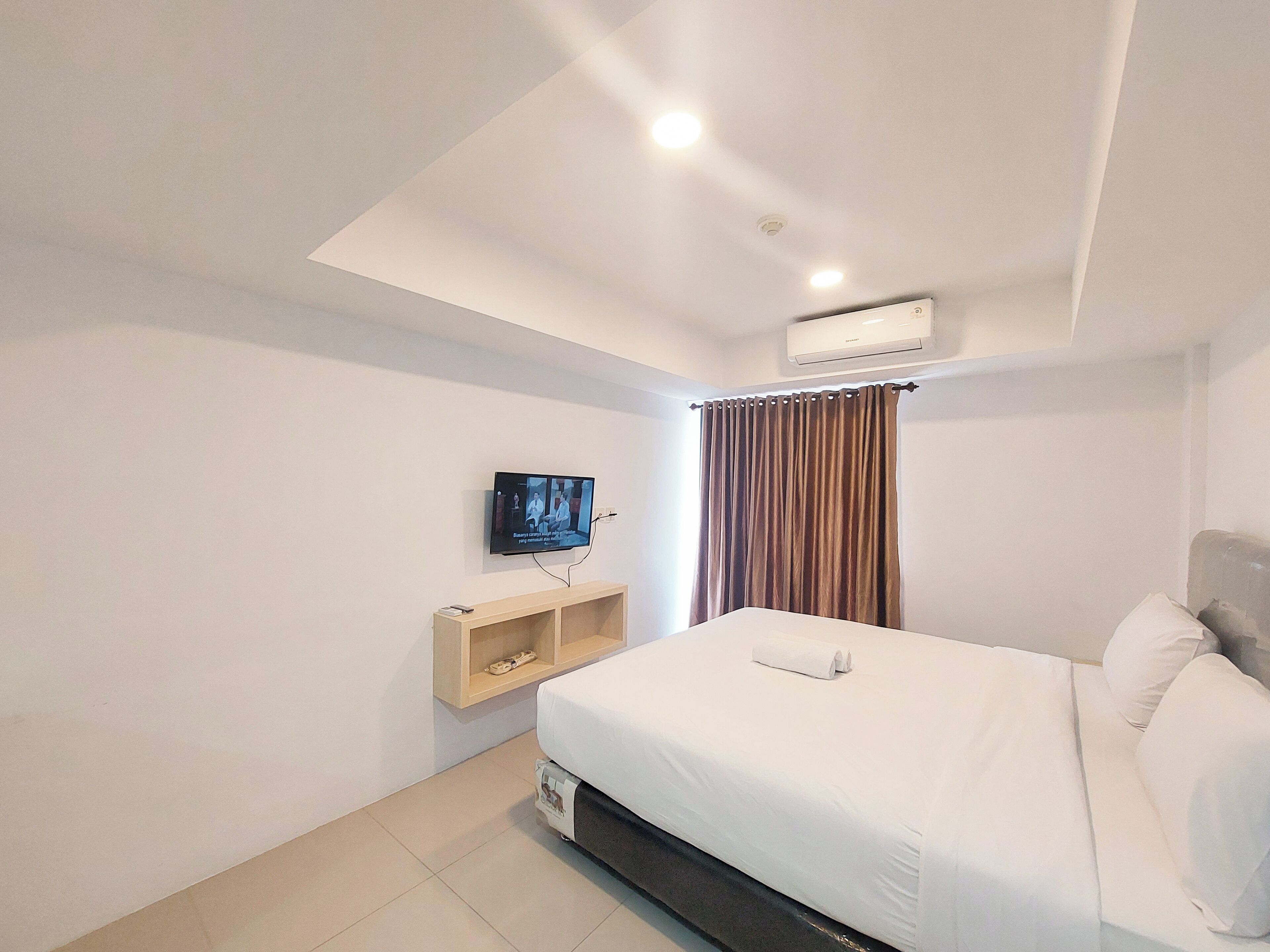 A hotel room with a bed, a TV mounted on the wall, a shelf with a remote control, and an air conditioning unit.
