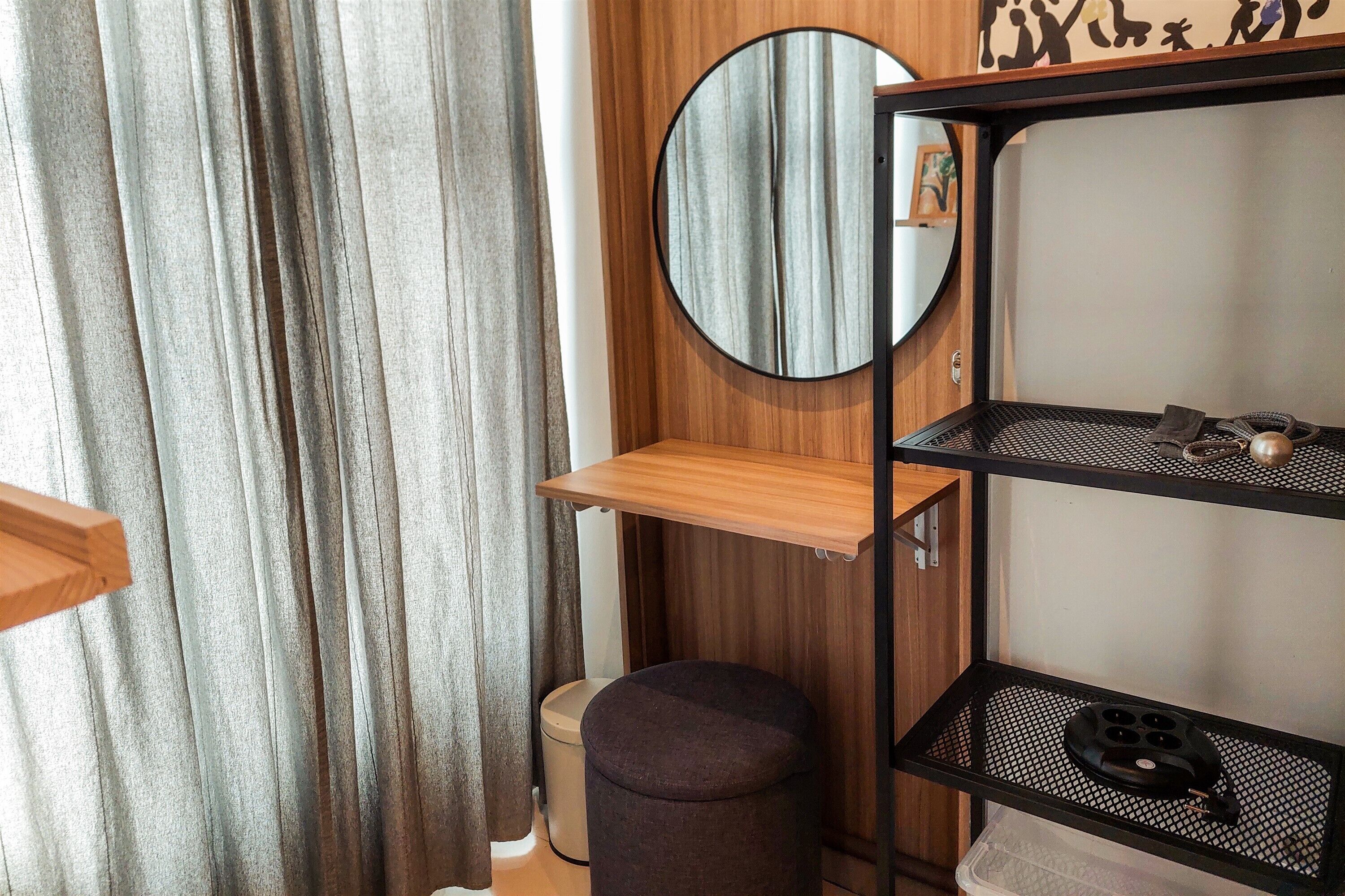 Una habitación con estante de madera, espejo redondo, taburete pequeño y un cubo de basura.