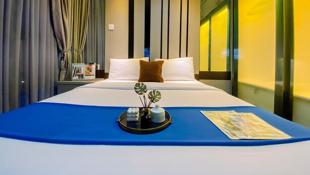 A modern hotel room with a large bed, a blue accent wall, a decorative tray with flowers, and a window with yellow lighting. - Homey 2Br At Gp Plaza Apartment (Jakarta)