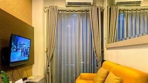 A modern living room with a flat-screen TV mounted on the wall, a sofa with cushions, a potted plant, and a window with curtains. - Homey 2Br At Gp Plaza Apartment (Jakarta)