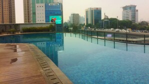 A swimming pool with a wooden deck, surrounded by tall buildings and a busy road. - Homey 2Br At Gp Plaza Apartment (Jakarta)