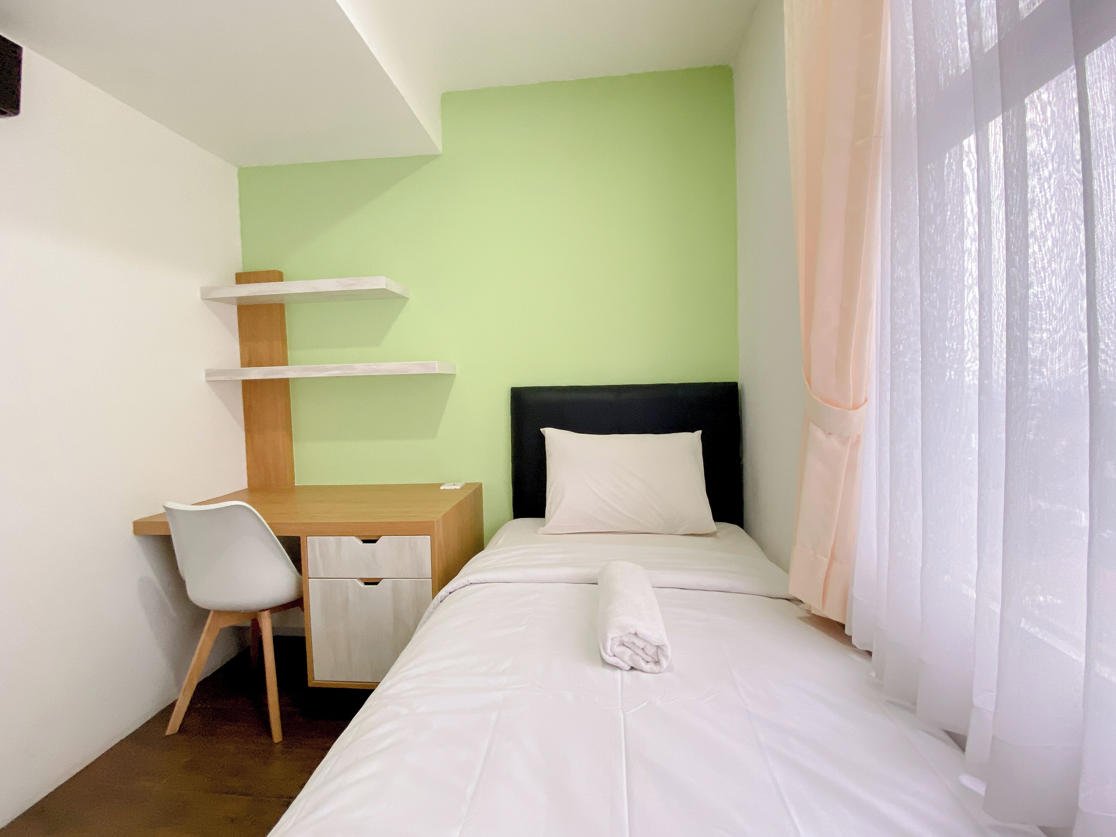 Una habitación de hotel con una cama, un escritorio con silla, una cómoda pequeña y una ventana con cortinas traslúcidas.