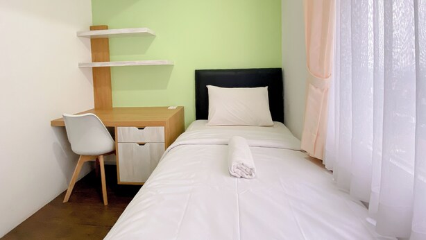 Una habitación de hotel con una cama, un escritorio con silla, una cómoda pequeña y una ventana con cortinas traslúcidas.