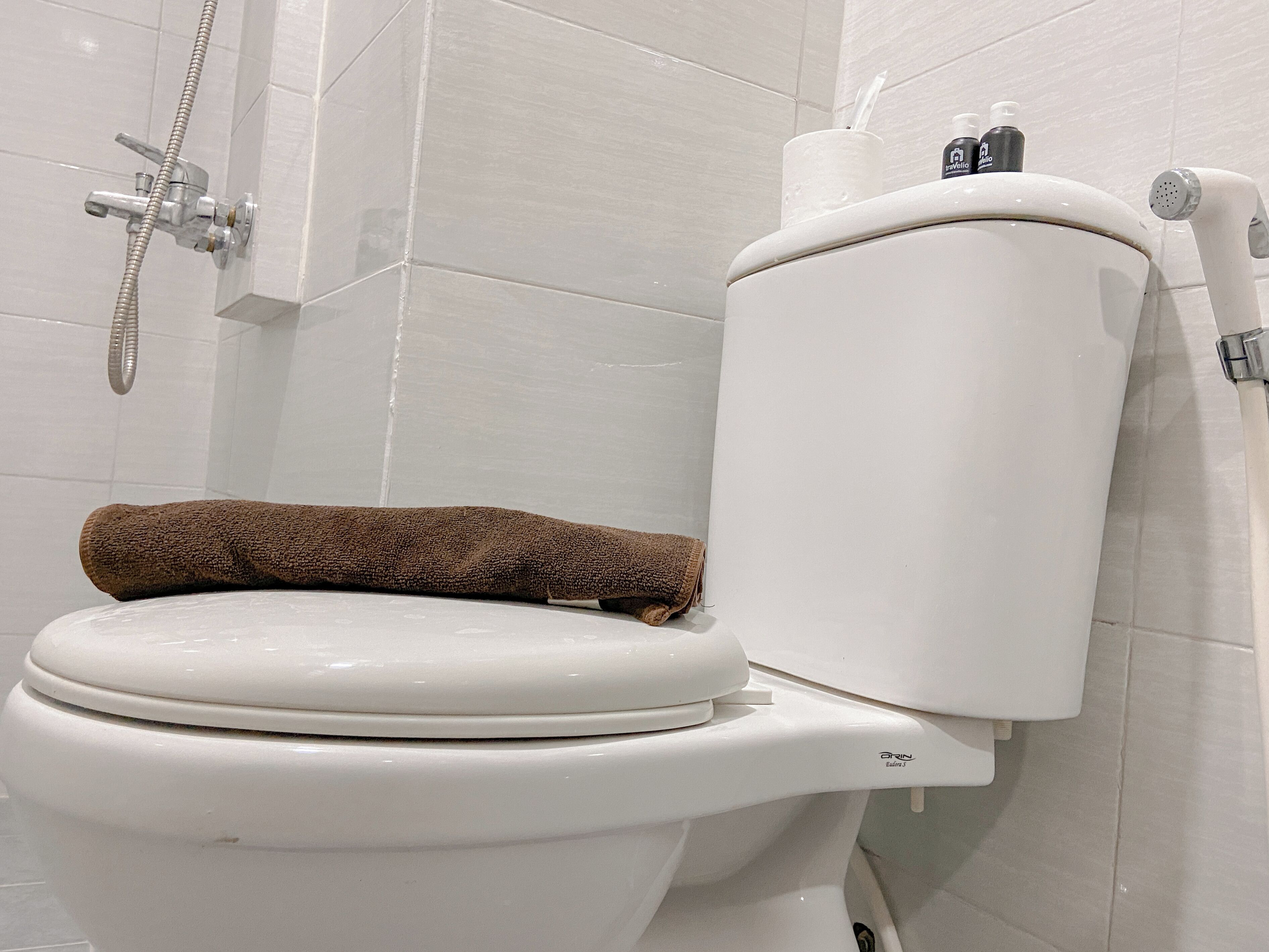 A white toilet with a brown towel on the seat and a showerhead mounted on the wall.