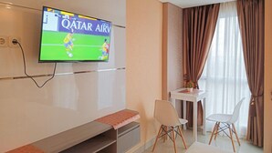 A hotel room with a flat-screen TV mounted on the wall, a small table with two chairs, a bench, and a window with curtains.
