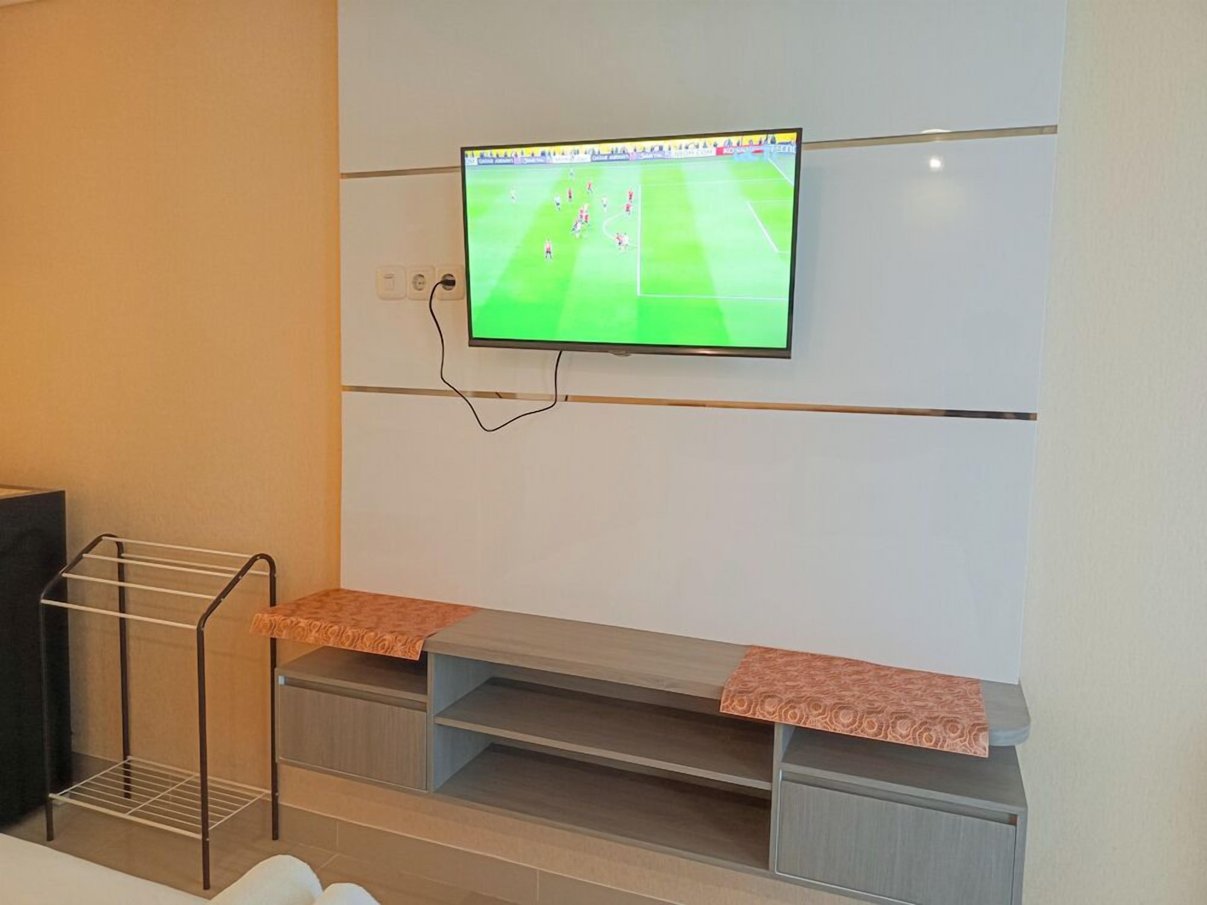 A room with a mounted TV displaying a soccer game, a metal drying rack, and a wooden bench with a patterned cover.