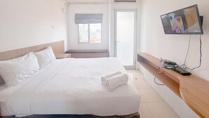 A hotel room with a bed, a TV mounted on the wall, and a window with a view of buildings. - Compact And Restful Studio At Pavilion Permata Apartment (Surabaya)