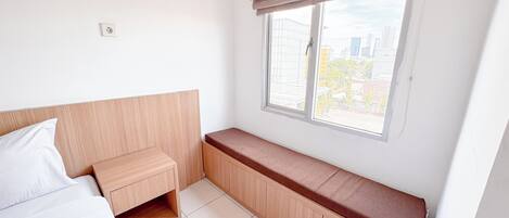A small, bright room with a bed, a wooden bench, and a window with a view of buildings.