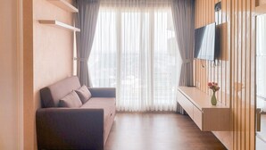 A modern living room with a sofa, a flat-screen TV mounted on a wooden panel, and a large window with sheer curtains.
