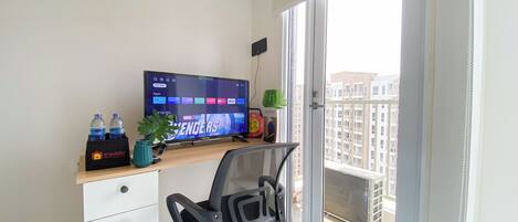 A small office space with a desk, a computer, a chair, a potted plant, and a view of a residential building through a sliding door.