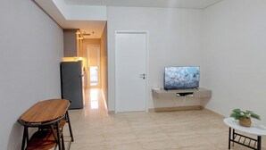 A modern living room with a television, a small table with a plant, a wooden chair, and a refrigerator. - Tranquil Space 1Br Apartment At Podomoro City Deli Medan (Medan)