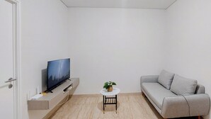 A modern living room with a flat-screen TV, a grey sofa, a small round table with a plant, and a white door. - Tranquil Space 1Br Apartment At Podomoro City Deli Medan (Medan)