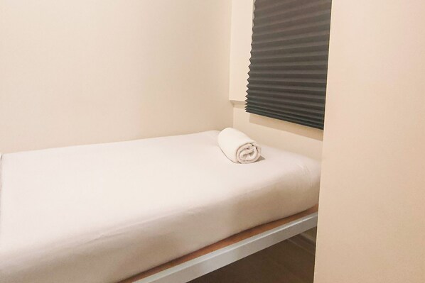 Una cama individual con una manta blanca enrollada cuidadosamente en una habitación pequeña y sencilla, con paredes lisas y una ventana con persianas.