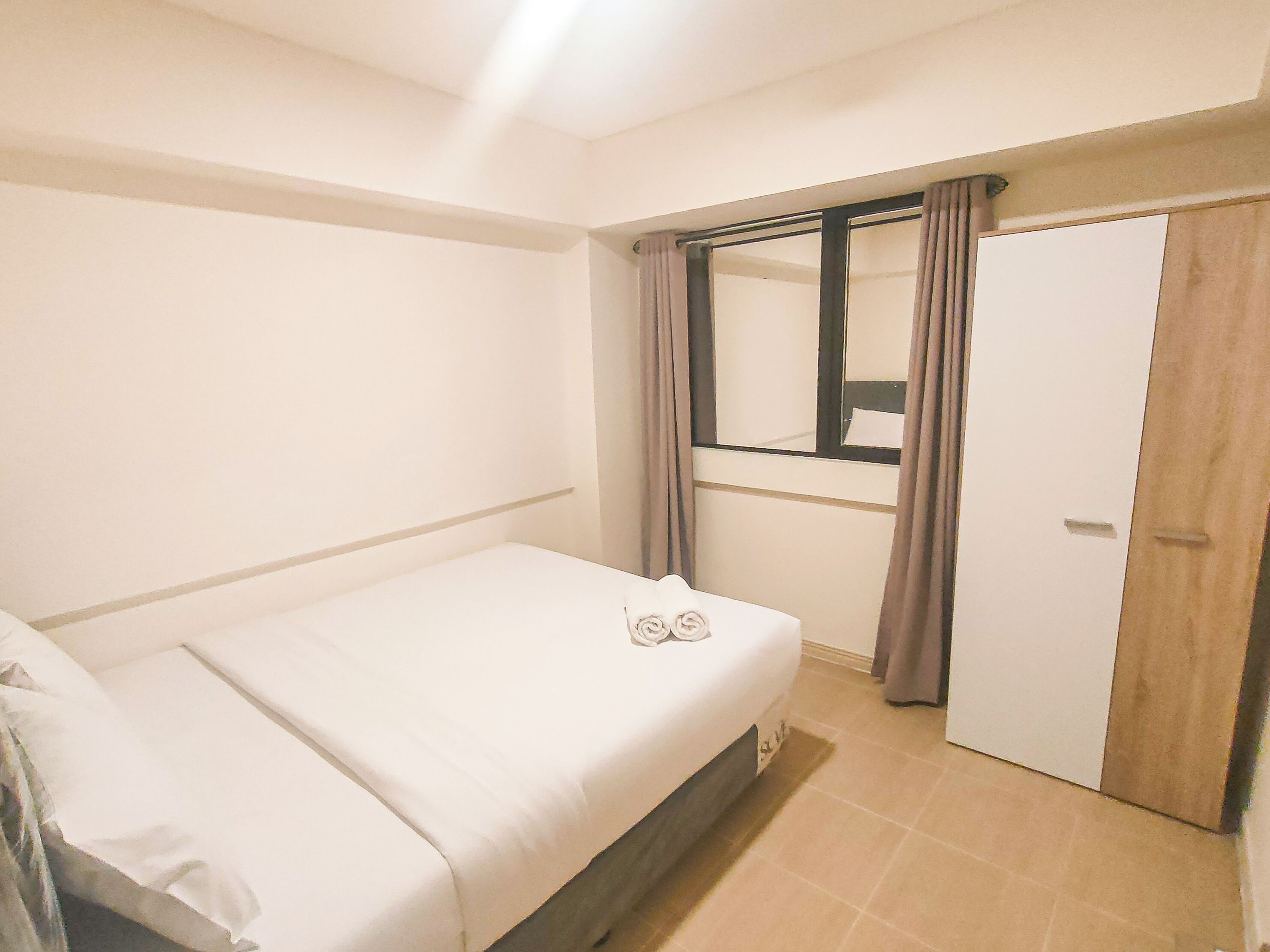 A small, clean room with a single bed, a wooden door, and a window with curtains.