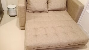 A beige sofa with two pillows and a trash can in a room with white walls and a window with blinds. - Marvelous And Cozy 2Br At Meikarta Apartment (Cikarang)